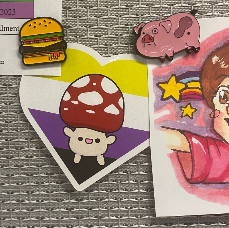 a sticker with a mushroom in front of the nonbinary flag, a hamburger enamel pin, an enamel pin of Waddles from Gravity Falls, and part of fan-made drawing of Mabel Pines.